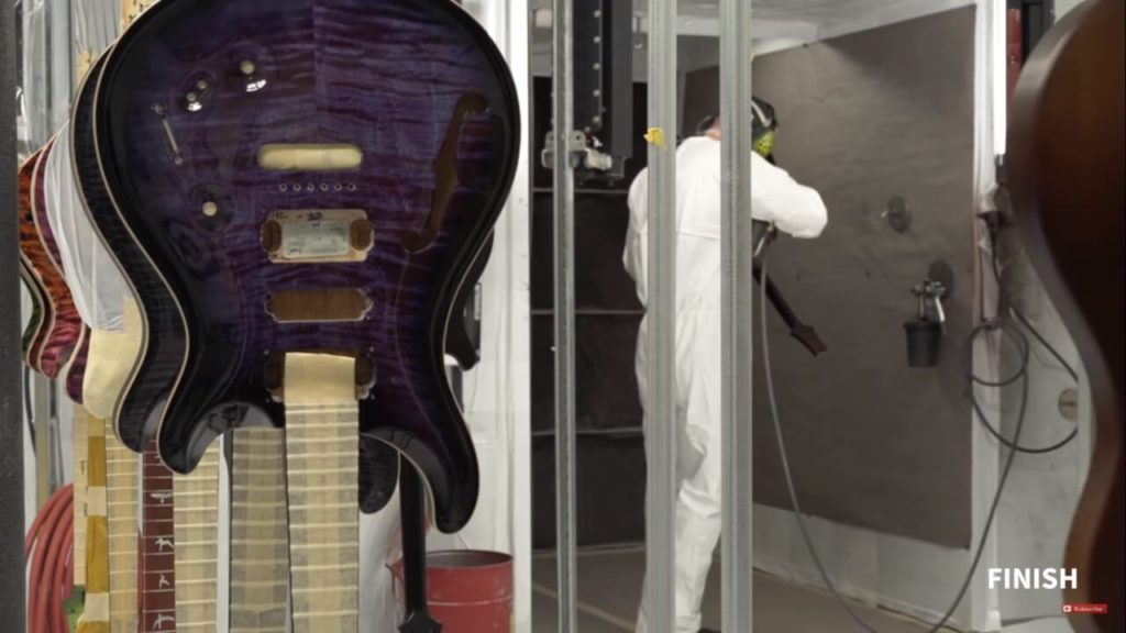Guitarras en proceso de secado y curado, atrás operario pinta una guitarra.